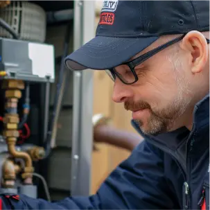 A man wearing glasses and a cap works on air conditioning machinery with pipes and wires, appearing focused on a maintenance or repair task.