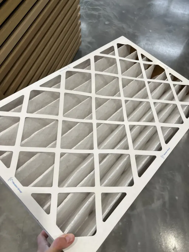 White diamond-pattern air filter panel being held, with stacked brown slatted panels in the background on a concrete floor