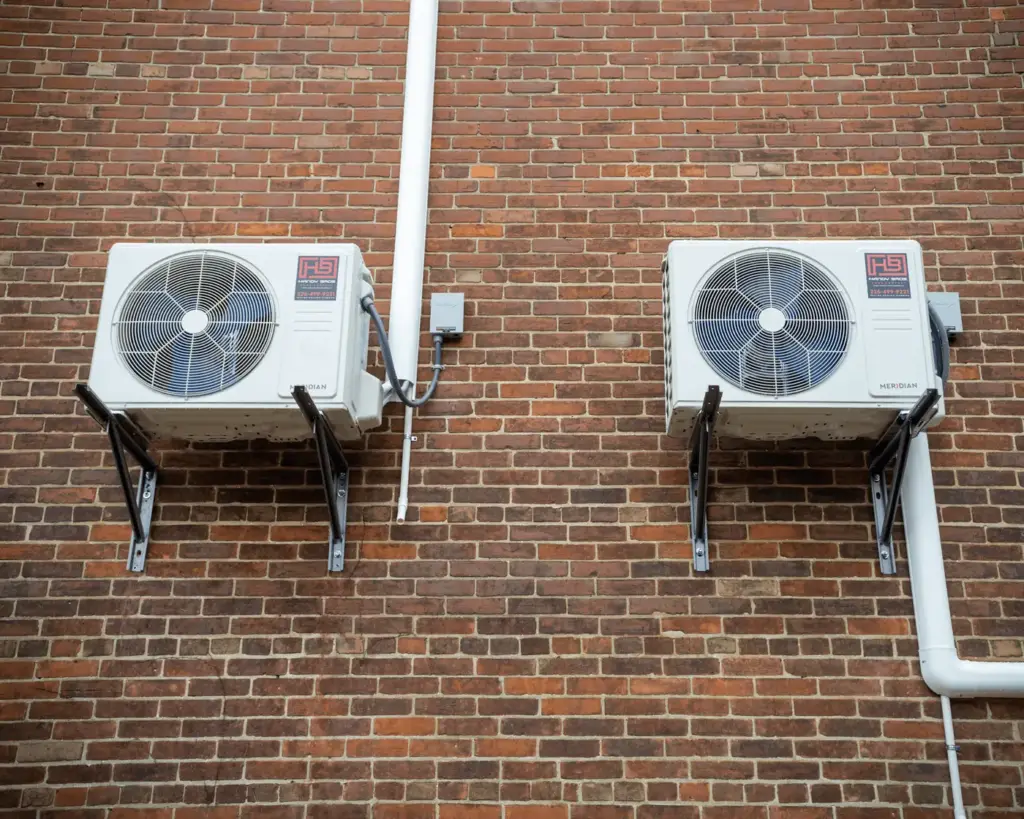 Two outdoor air conditioning condensers mounted on a brick wall with a white drain pipe between them and metal supports beneath each unit.