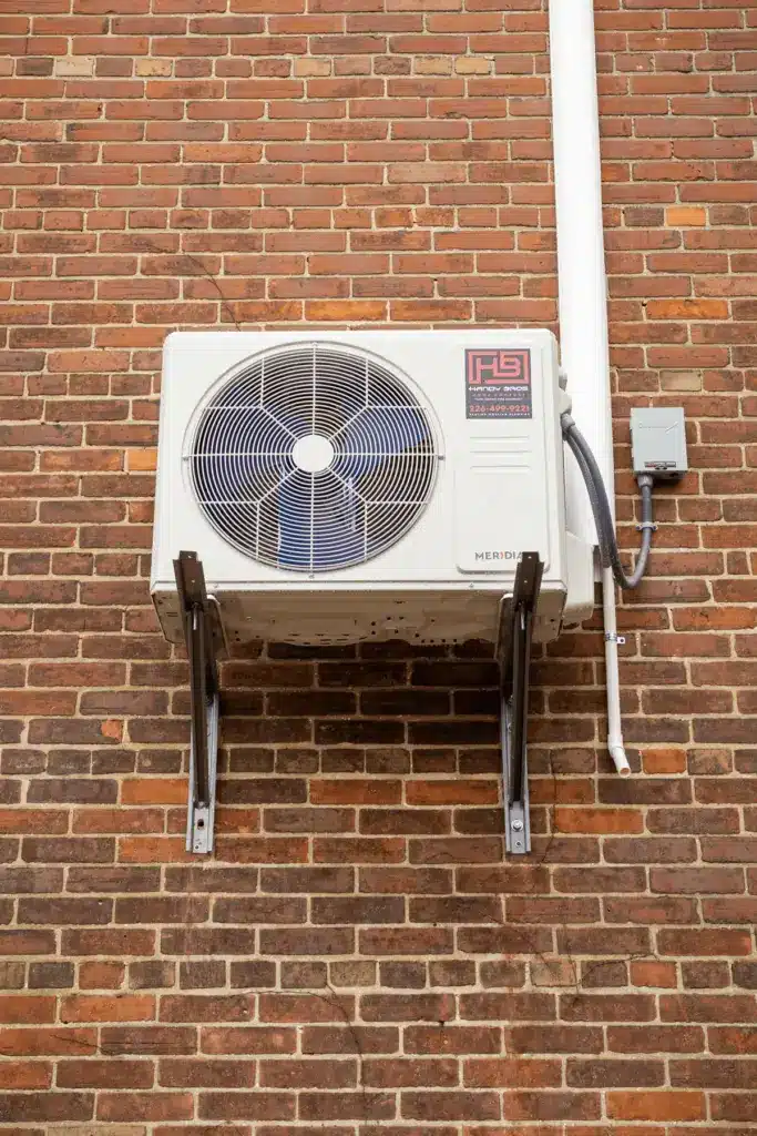 Outdoor air conditioner condenser unit mounted on a brick wall with metal brackets and connected piping.