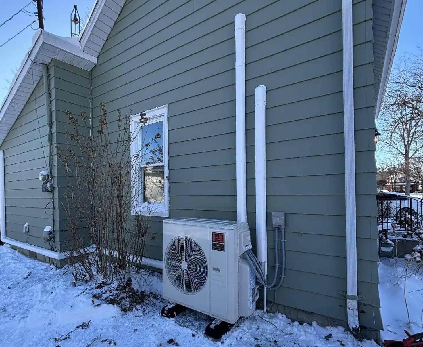 Exterior of a green house with an outdoor air conditioner unit and white vent pipes on a snowy day, by a small leafless shrub.