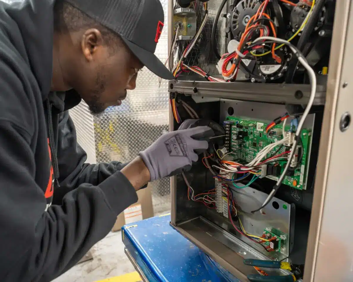 Technician wearing gloves and cap inspecting exposed circuit boards and wiring inside an open cabinet of a machine.