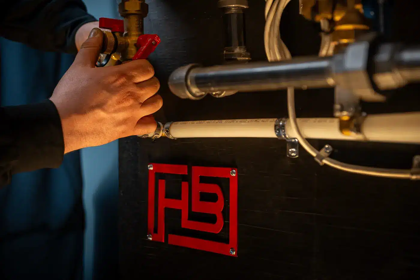 A close-up of a hand turning a red valve on an industrial pipe assembly mounted to a dark wall with a red logo plate nearby.