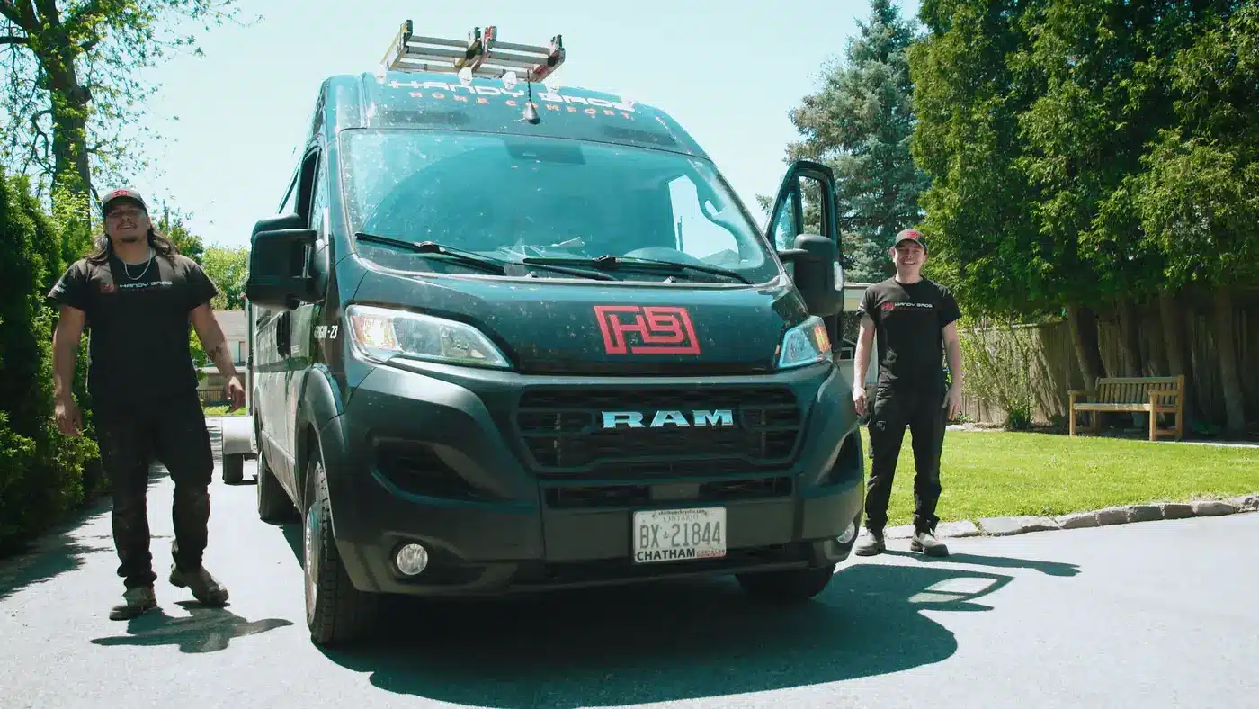 Two men stand beside a dark teal RAM work van in a residential driveway on a sunny day, with green lawn and trees in the background.