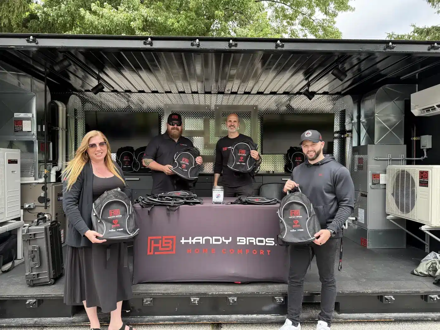 Four people stand inside a trailer showroom, each holding black Handy Bros backpacks with red logos on a table covered by a Handy Bros banner.