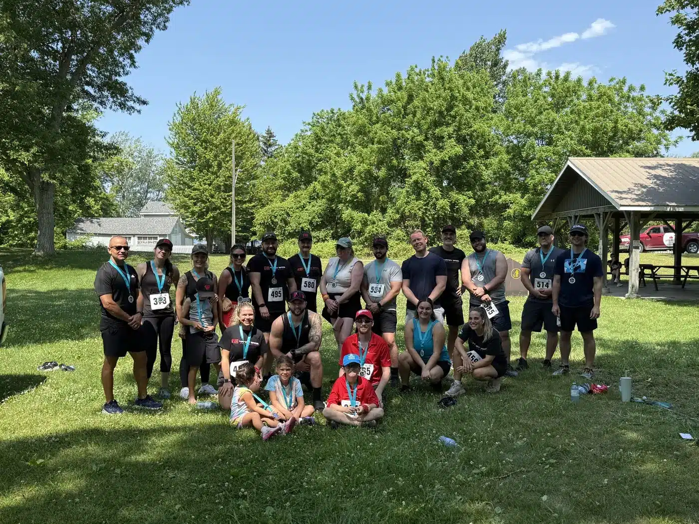 Group of runners with medals posing on a sunny park lawn after a race, trees and a pavilion in the background.