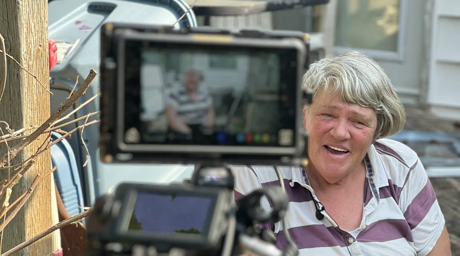 Older woman with short gray hair smiles while being filmed outdoors; a camera monitor is visible in the foreground.