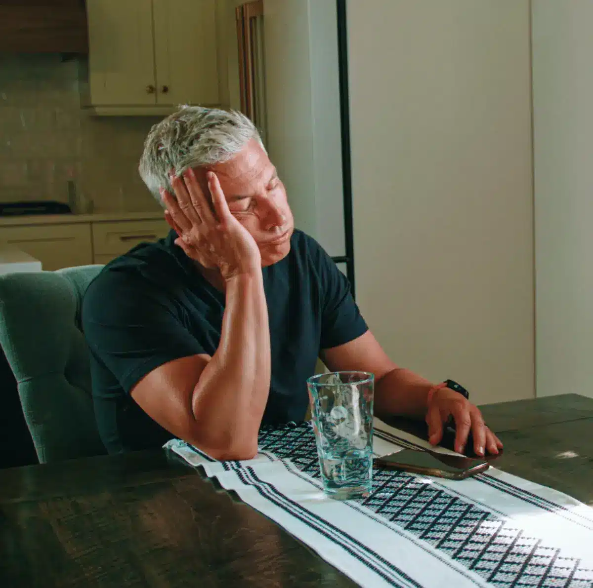 Man with gray hair sits at a dining table, resting his head on his hand, looking fatigued or asleep.