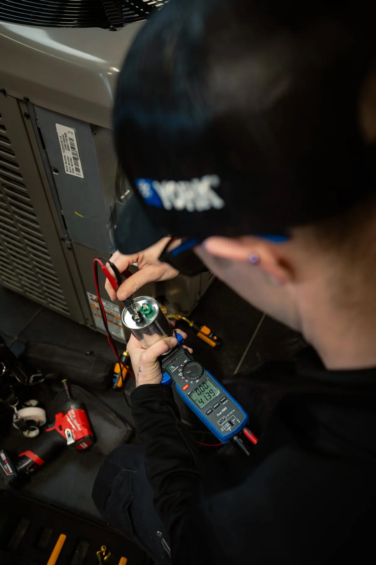 Technician wearing safety glasses uses a digital multimeter to test a capacitor inside an HVAC unit.