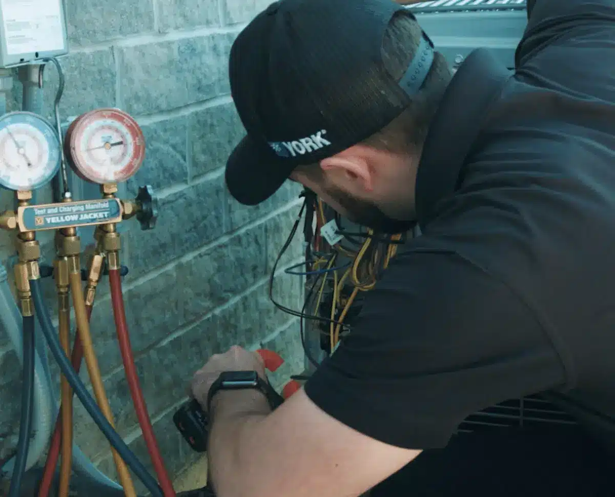 HVAC technician crouching beside a refrigerant manifold with colorful hoses connected to a wall-mounted system outside.