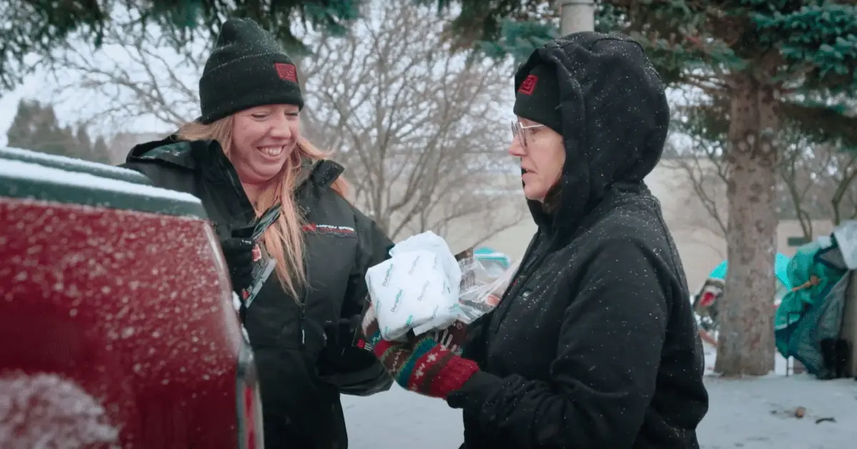 Two women in winter gear exchange a bag near a red car in a snowy outdoor setting, smiling.