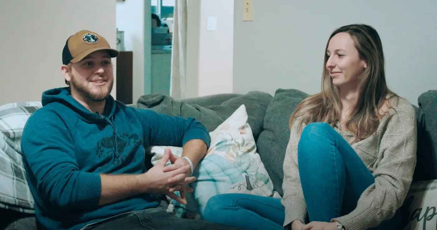 Two people sit on a gray sofa in a living room, chatting casually; man in a teal hoodie and cap, woman in a beige sweater and jeans, smiling.