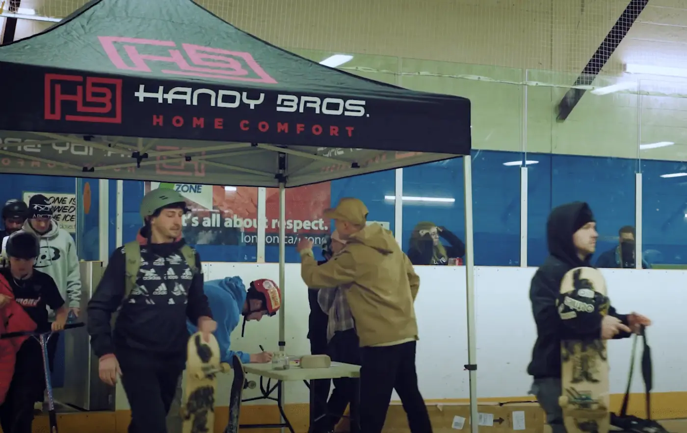 Skatepark scene under a green Handy Bros. tent with people wearing helmets and holding skateboards indoors.