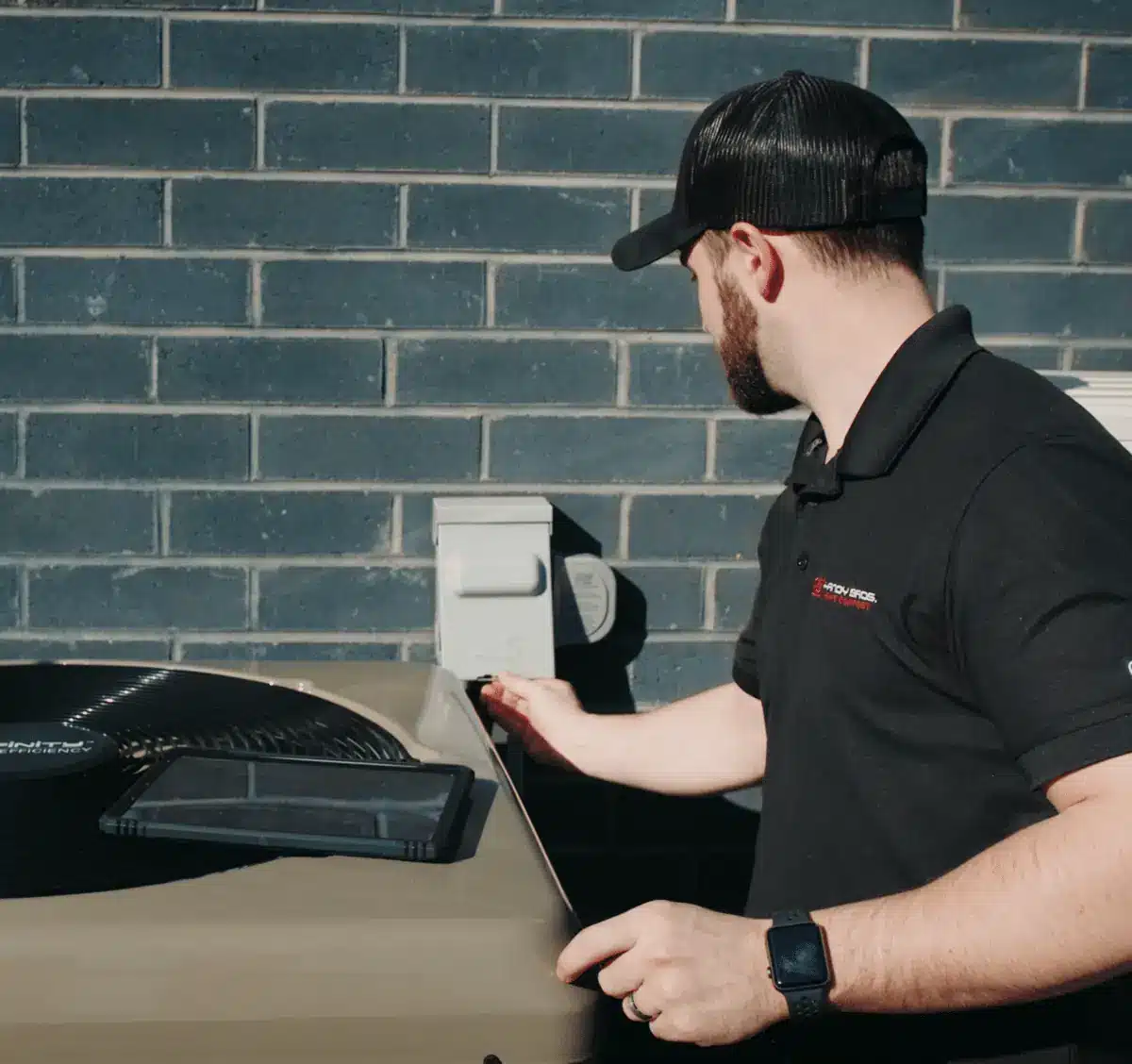 Technician in a black polo and cap services an outdoor HVAC condenser against a blue brick wall, with a tablet resting on the unit nearby.