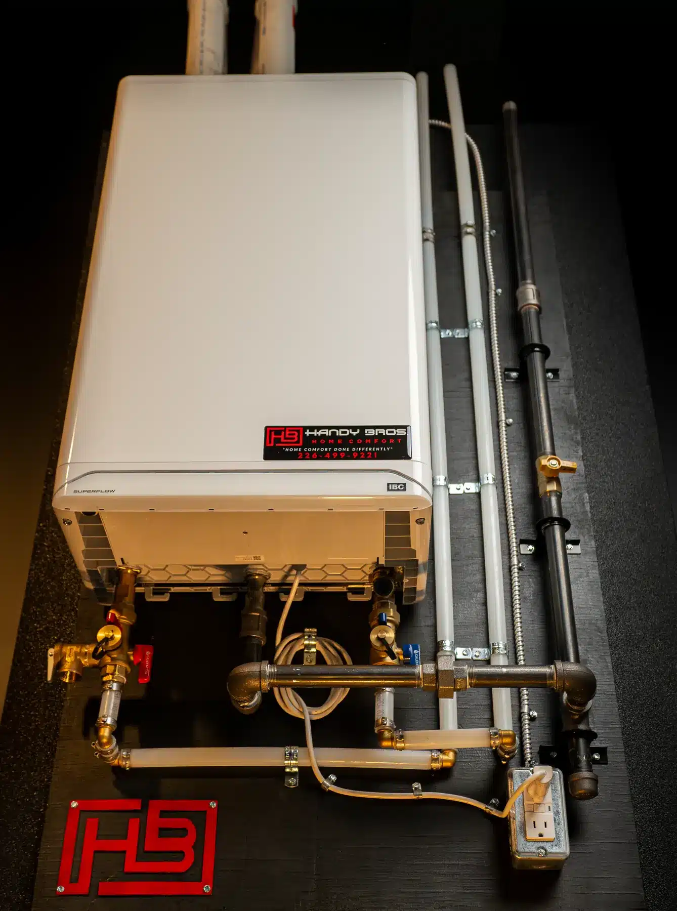 Wall-mounted white boiler with multiple pipes, valves, and an electrical outlet; red HB Handy Bros sign visible below the unit.