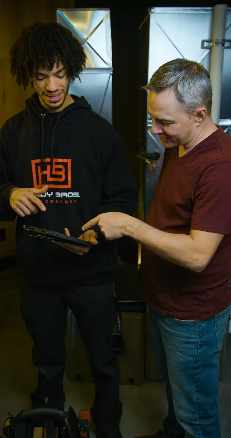 Two men stand in a workshop looking at a tablet, one pointing at the screen as they discuss it.