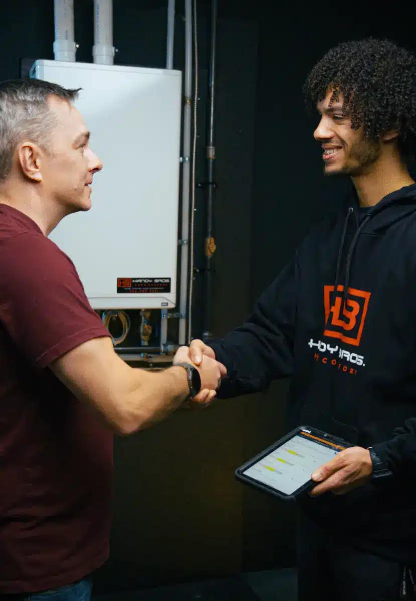 Two men shake hands in a technical setting beside electrical equipment, one holding a tablet.