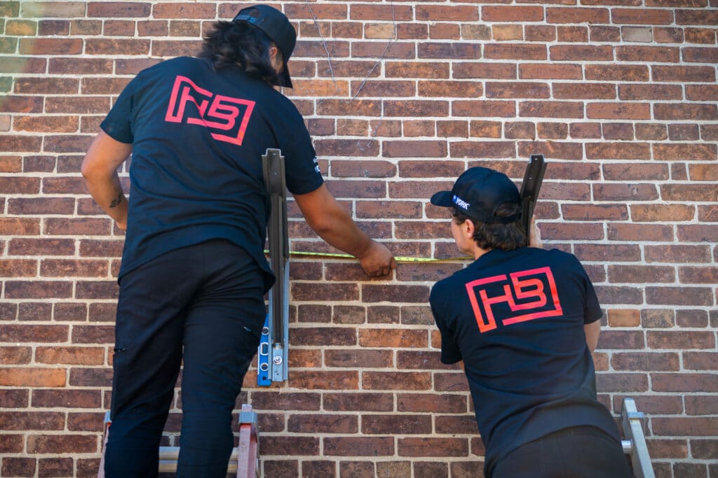 Two workers on ladders measure a brick wall with a tape measure, wearing matching black shirts with pink logo on the back.