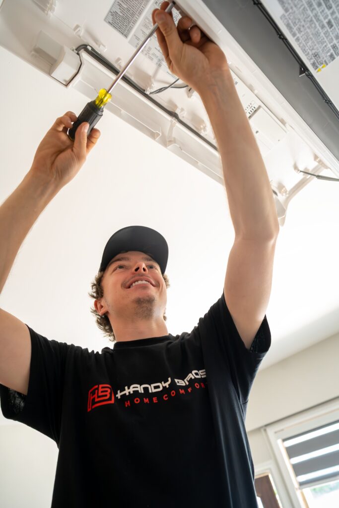 Technician in a black shirt and cap using a screwdriver to work on a ceiling-mounted appliance (AC unit) above him.