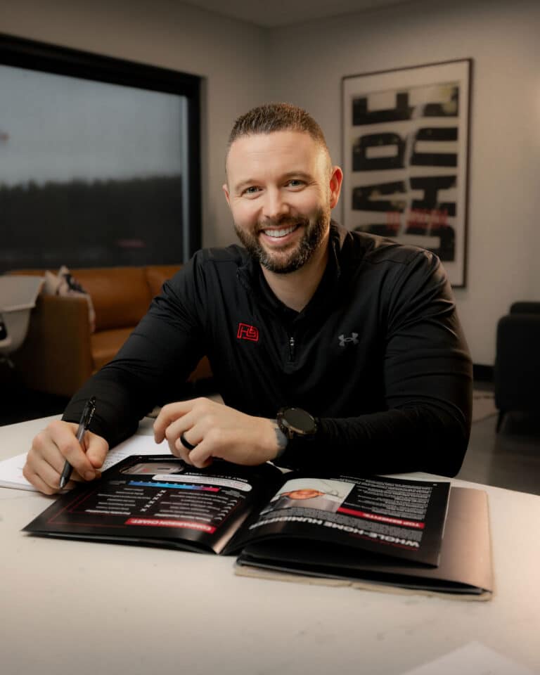 Smiling man with a beard signs a magazine at a white table in a casual office setting.